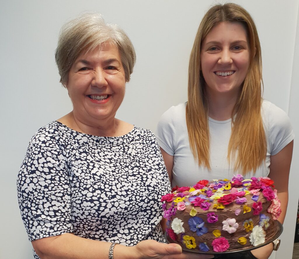 Lucia Forman and Deanne Gibbs (Advocate) holding Lucia's farewell cake. A chocolate cake covered in colourful flowers.
